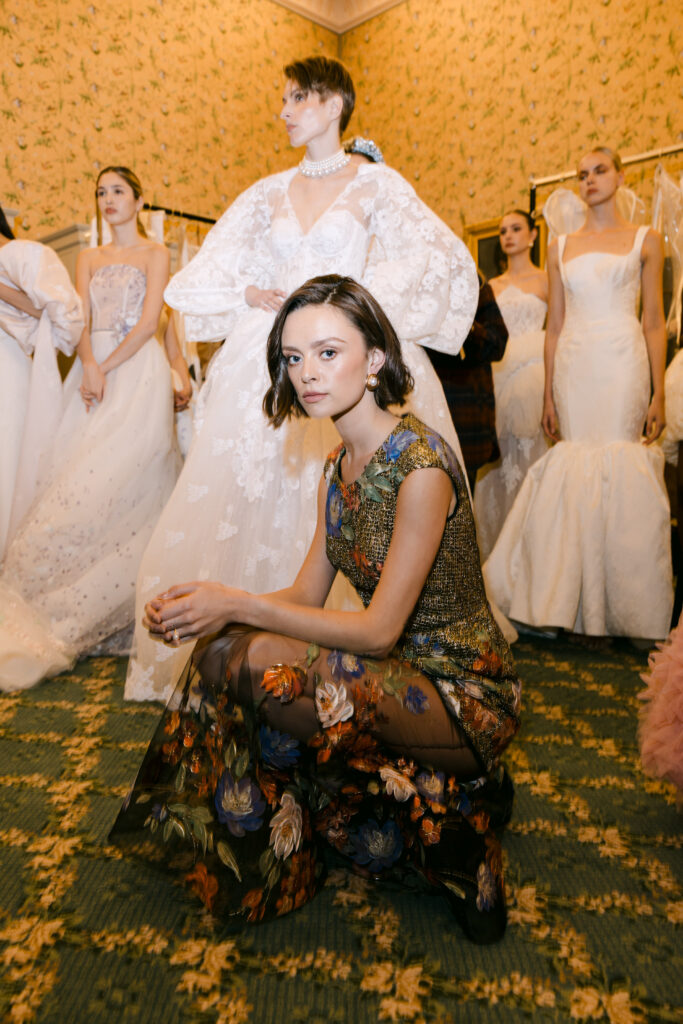 A woman in a floral dress crouches in front of a group of models wearing elegant white bridal gowns, standing in a room with patterned wallpaper and green carpet.
