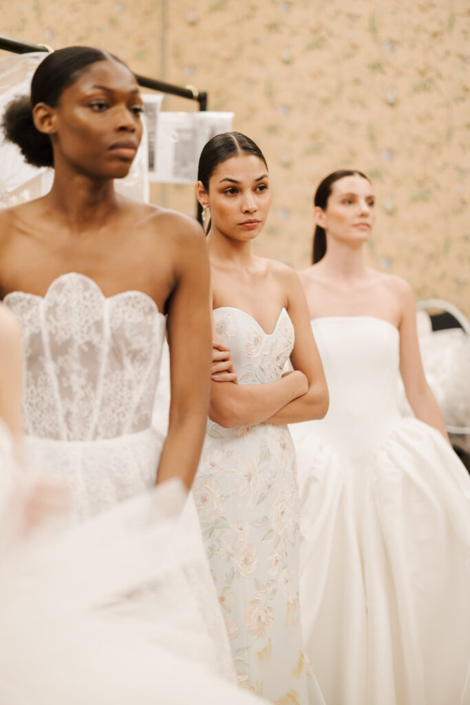 Three women in elegant, strapless wedding dresses stand together indoors, looking thoughtfully ahead. The background features light, patterned wallpaper, and bridal gowns are visible in soft focus.