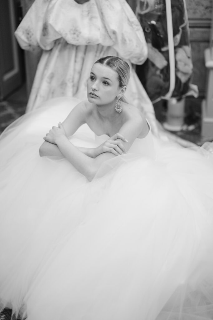 A woman in a voluminous white gown sits with her arms crossed over her knees, looking thoughtfully upward; her hair is slicked back and she wears large earrings. The image is in black and white.