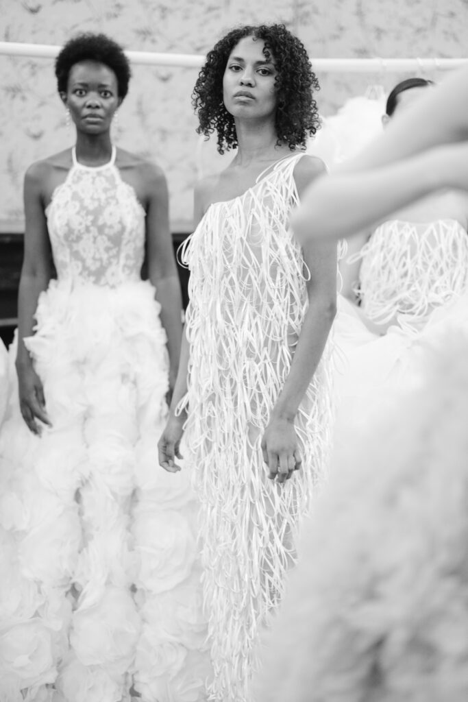 Two women in elaborate white gowns stand side by side; one wears a dress with floral textures, the other sports a dress with long, string-like details. Both look toward the camera in a softly lit, elegant setting.