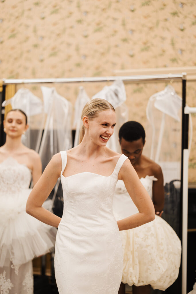 A smiling bride in a white wedding dress stands with her hands on her hips. Two other women in white dresses are in the background, with a clothing rack and beige floral wallpaper behind them.