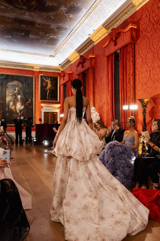 A model with long braids walks a runway in a grand, ornate room wearing a strapless, flowing white gown with a floral pattern. The audience watches and takes photos as she passes by.