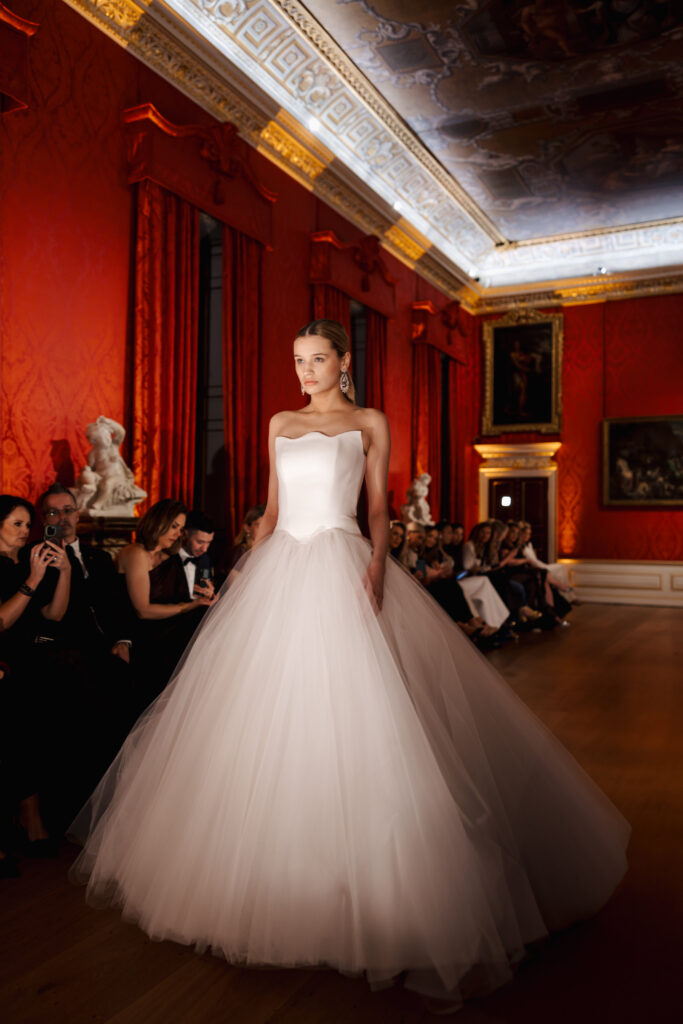 A model in a strapless, full-skirted white wedding gown walks down a runway in an ornate room with red walls, gold accents, statues, and a painted ceiling. Seated guests watch and take photos.