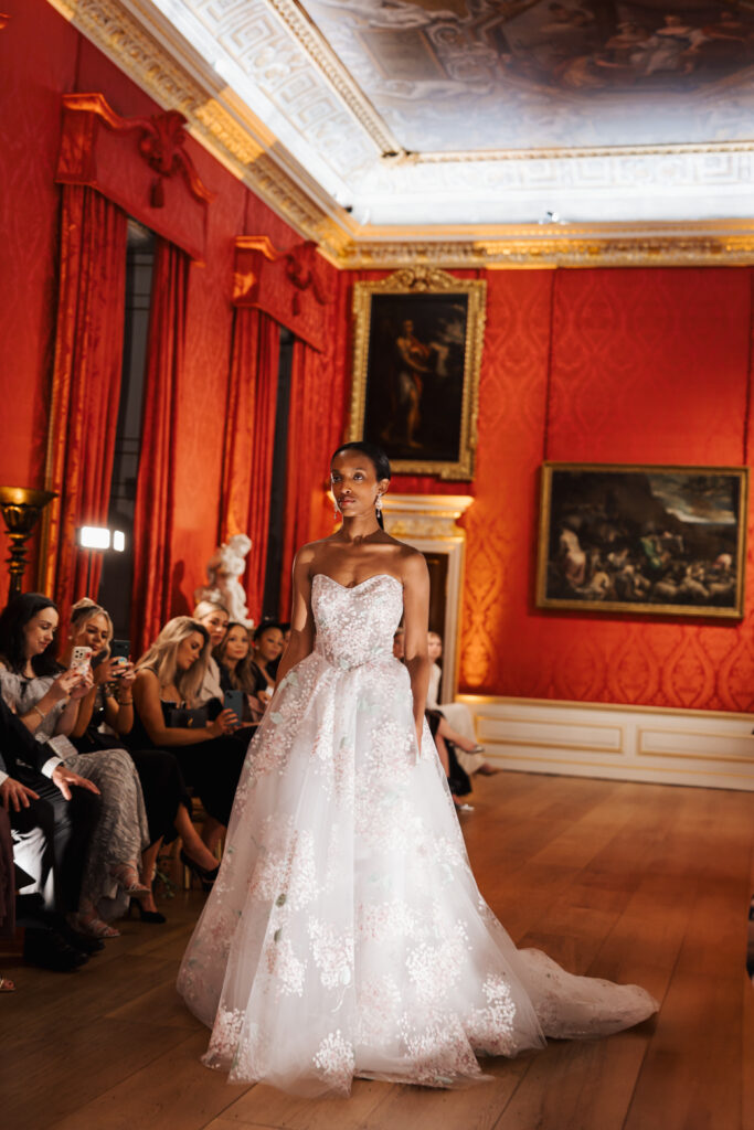 A model walks down a wooden runway in an ornate red room, wearing a strapless white ball gown with floral lace details. Seated guests watch, and classic paintings and sculptures adorn the walls.