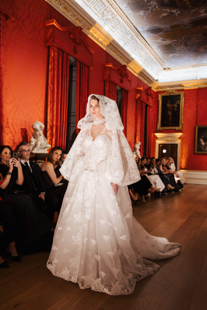 A model walks a runway in an ornate room, wearing an elaborate white lace wedding dress with a large hooded veil. Seated guests watch on either side; the walls are red with gold accents and classical paintings.