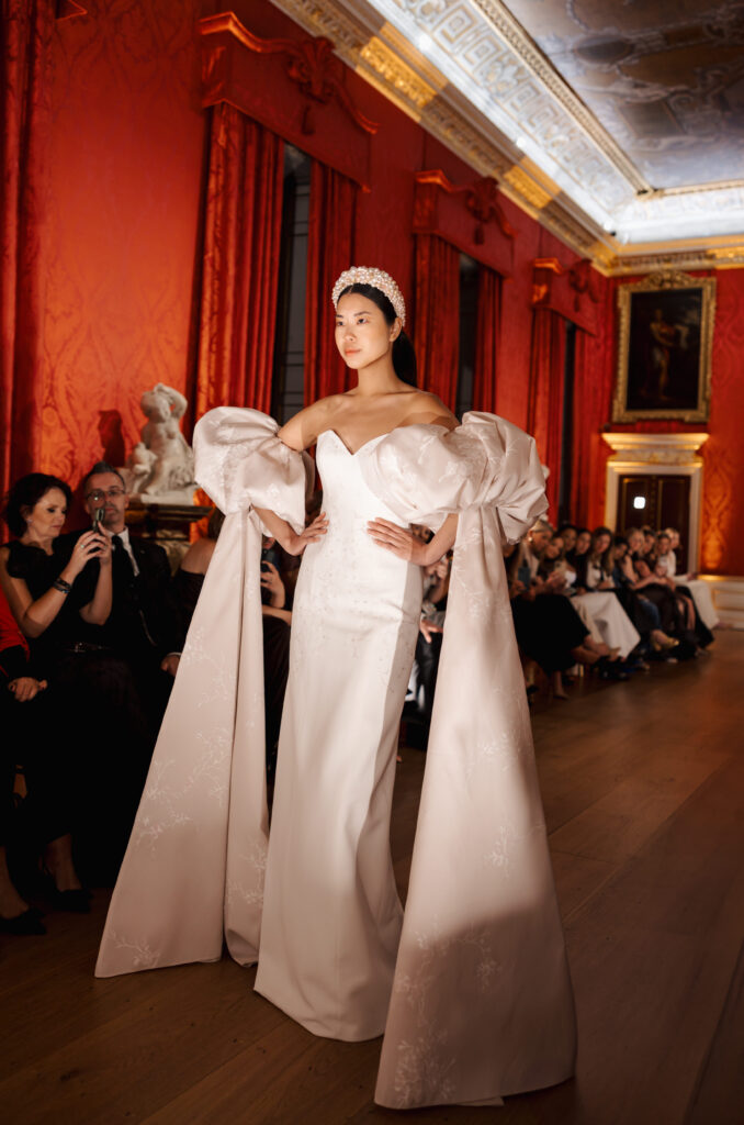 A model walks a runway in a white gown with dramatic puffed sleeves and long, flowing fabric, wearing a pearl headpiece. The fashion show is set in an ornate, red-walled room with classical decor and seated audience.