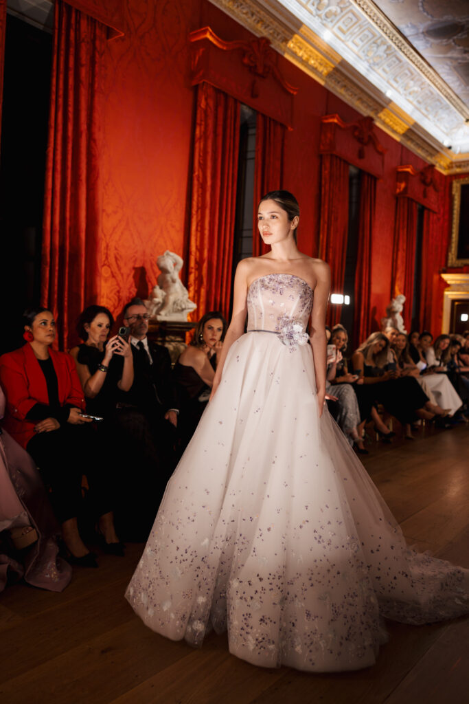A model in a strapless, pale lavender ball gown with sparkling details walks a runway in an ornate room with red walls, tall curtains, and an audience seated along the sides.