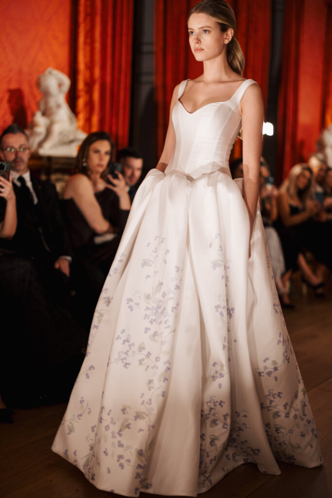 A model walks a runway wearing a white, full-skirted gown with delicate lavender floral patterns. The audience watches in an elegant setting with red drapes and classical sculptures in the background.