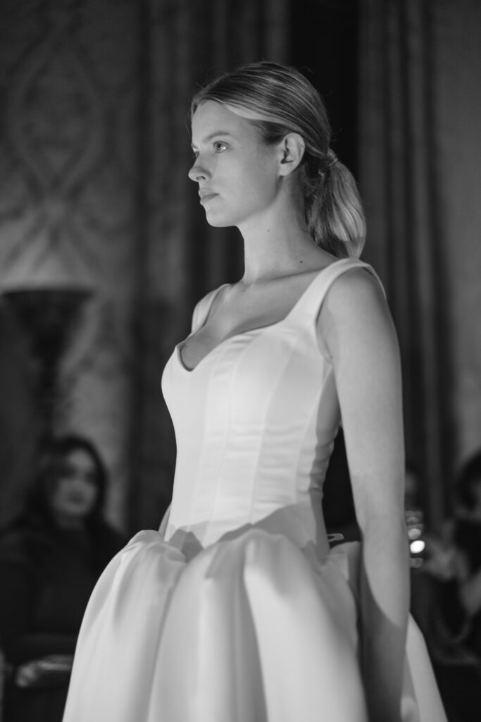 A woman with light hair in a low ponytail wears a sleeveless white dress with a fitted bodice and voluminous skirt, standing in profile under soft lighting at a formal indoor event. The image is in black and white.