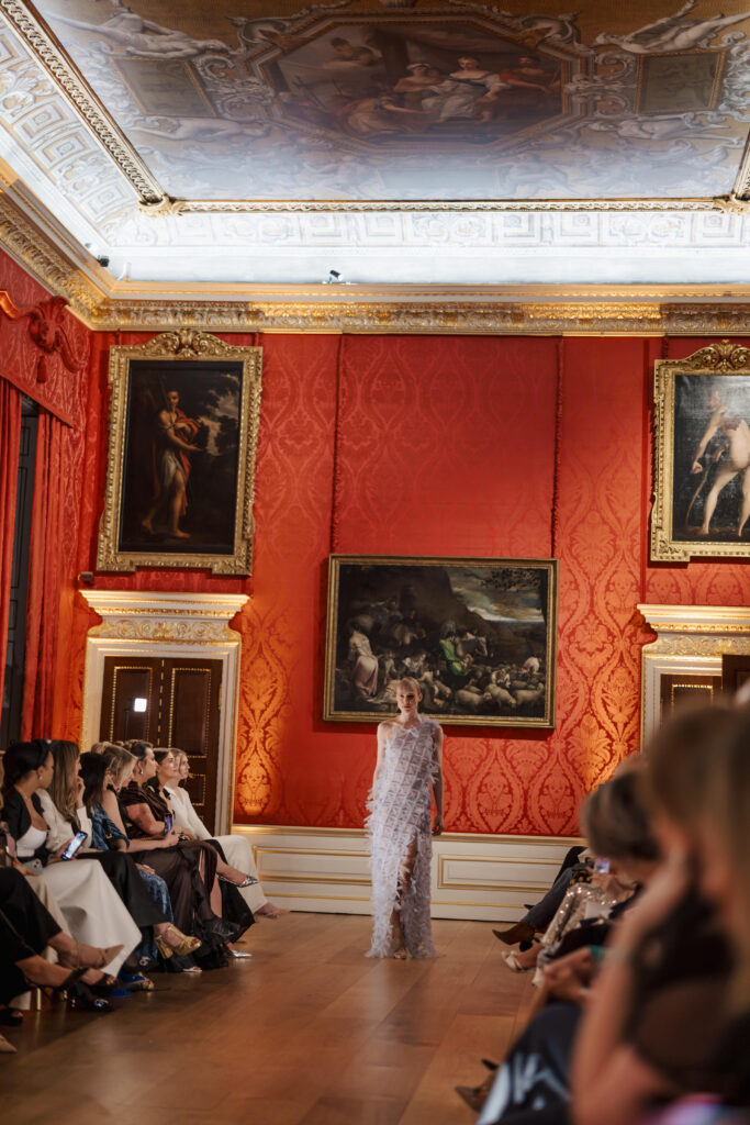 A model in a sheer, embellished gown walks down a runway in an ornate room with red walls, gold accents, and large classical paintings, while an audience sits on either side.