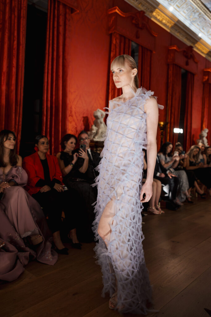 A model walks a runway in a sheer, geometric-textured dress, while an audience in elegant attire watches from both sides in a lavish room with red curtains and ornate decor.