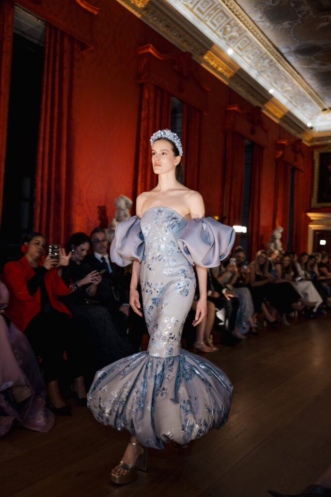 A model walks a runway in an ornate room, wearing a glamorous off-the-shoulder gown with dramatic puffed sleeves and a flared hem. The dress is silver-blue with floral patterns, and she wears a jeweled headpiece. Audience members watch and take photos.