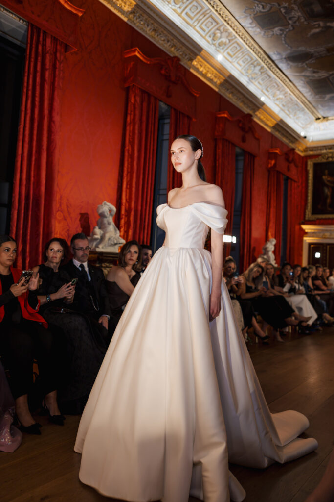 A model walks down the runway in an elegant off-the-shoulder white gown with a full skirt, as an audience seated in a grand, ornately decorated room with red walls and tall curtains looks on.