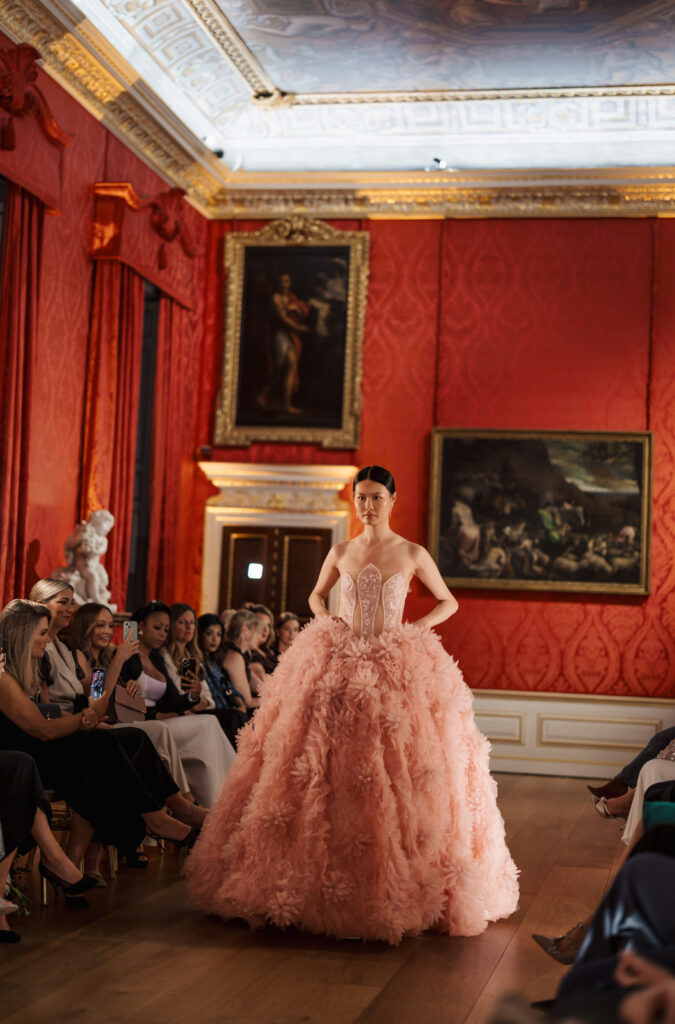 A model walks down a runway in an ornate room, wearing a strapless, voluminous pink gown with a feathered skirt. Seated guests line both sides of the runway, watching and taking photos. Paintings hang on the red, patterned walls.