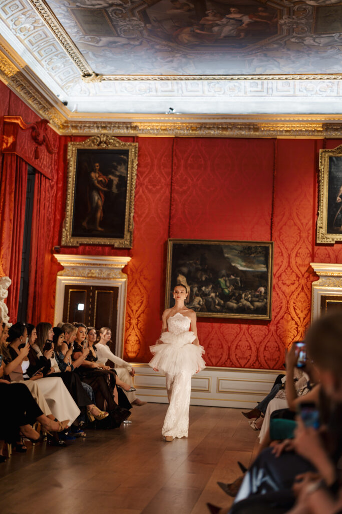 A model in a white, detailed gown walks down a runway in an ornate room with red walls, gold accents, classical paintings, and an audience seated on both sides.