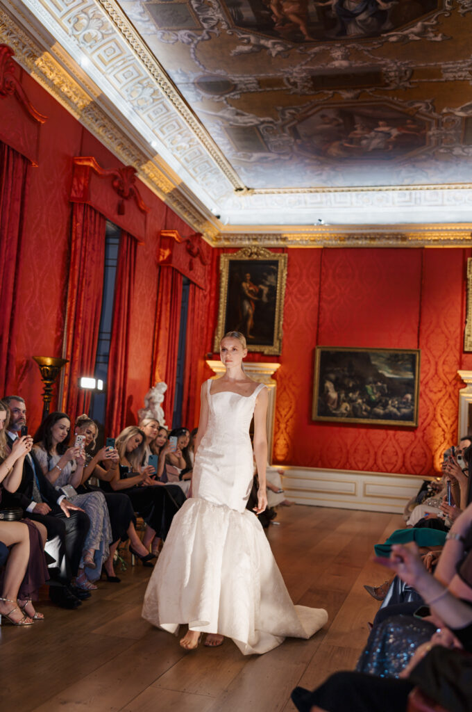 A model in a white, elegant gown walks down a runway in a lavish room with red walls, ornate ceiling, classical paintings, and an audience seated on both sides taking photos.