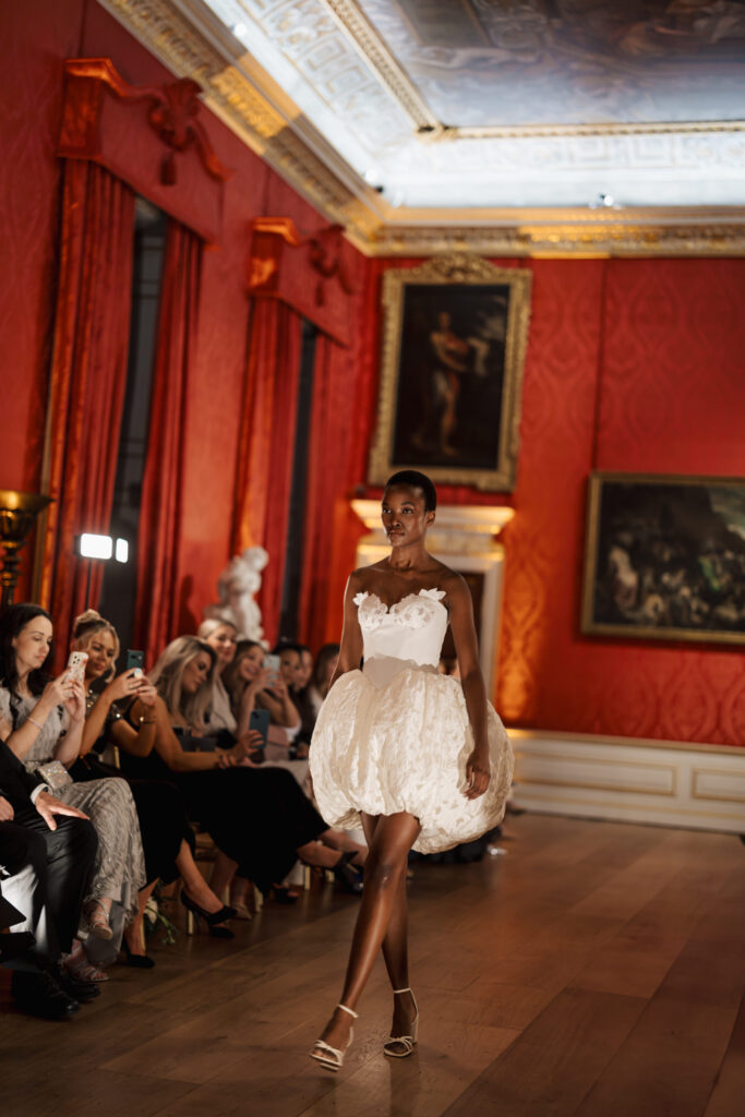 A model walks down a runway in an elegant room with red walls and classical paintings, wearing a white, floral-embellished, strapless dress with a voluminous skirt. Audience members seated nearby watch and take photos.