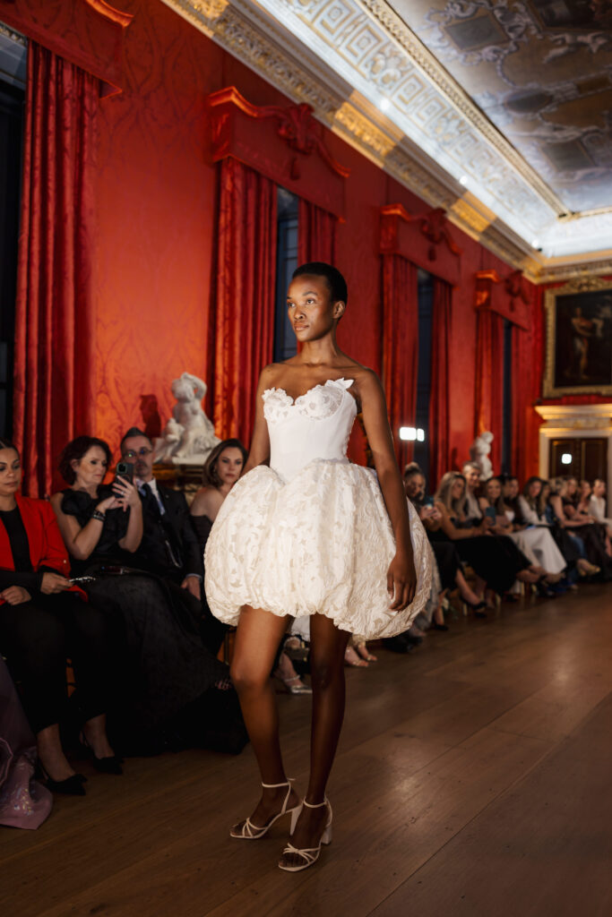 A model walks down a runway in an ornate room, wearing a white strapless dress with a voluminous, textured skirt and strappy heels, while an audience watches.