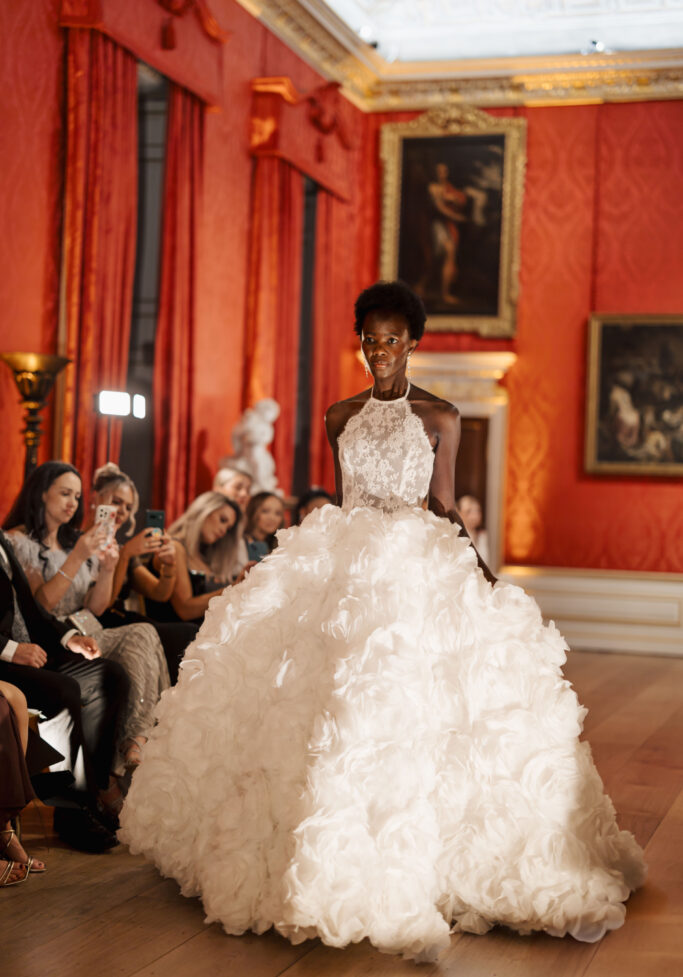 A model walks down a runway in an elegant white ball gown with a textured, voluminous skirt, as an audience seated on either side watches and takes photos in a lavish, red-walled room with classical paintings and sculptures.