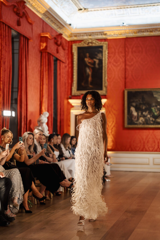 A model walks down a runway wearing a textured white dress in an ornate room with red walls, gold accents, framed paintings, and an audience seated along the sides.