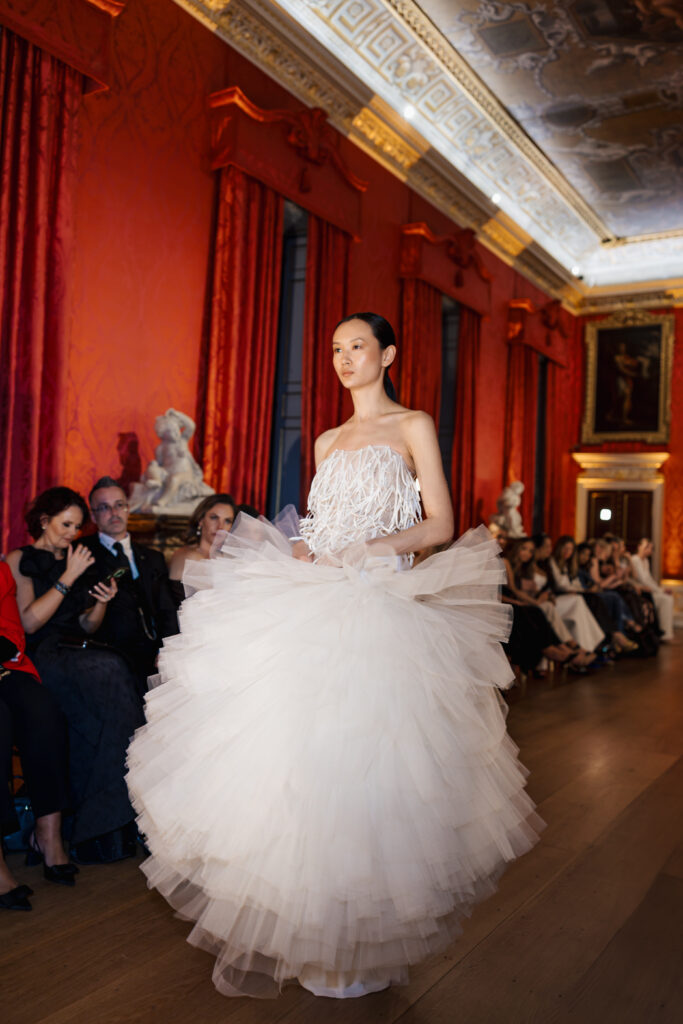 A model walks down a runway in an ornate room, wearing a voluminous white tulle gown with a detailed bodice, as an audience watches and takes photos. Red curtains and classical decor are visible in the background.