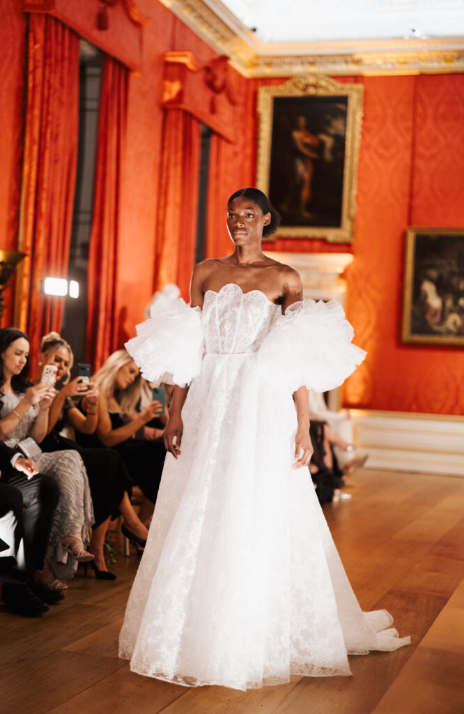 A model wearing an elegant white gown with voluminous tulle sleeves walks on a wooden runway in a richly decorated red and gold room, while an audience seated nearby takes photos.