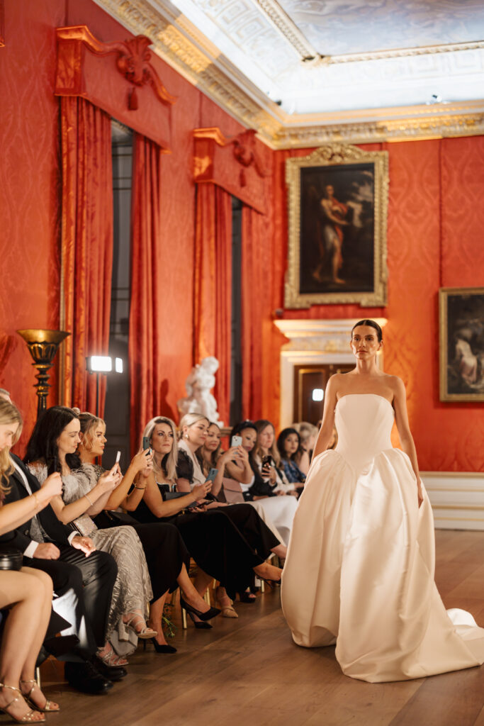 A model in a white strapless ball gown walks down a runway in an elegant room with red walls, ornate gold accents, and framed paintings. Seated guests watch and take photos with their phones.