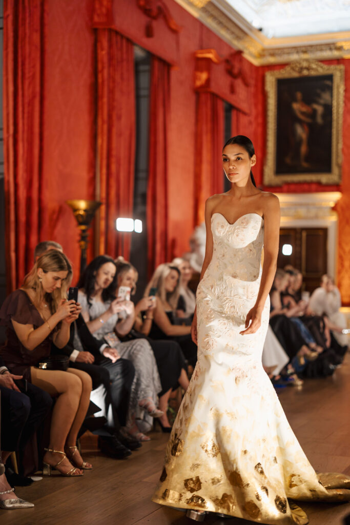 A model in a strapless white and gold gown walks down a luxurious runway with red walls, gold accents, and an audience seated on either side, some photographing the event.