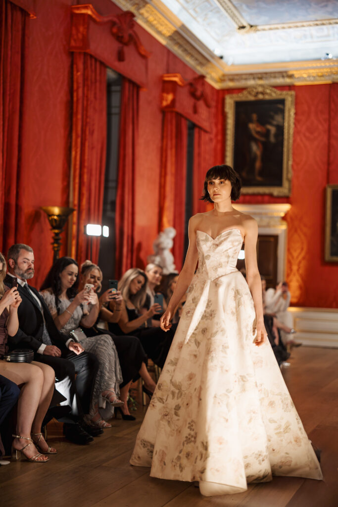 A model walks down a runway in an elegant, strapless, floral ball gown as an audience seated on the side watches and takes photos; the setting is a grand, ornate room with red walls, gold trim, and large framed paintings.