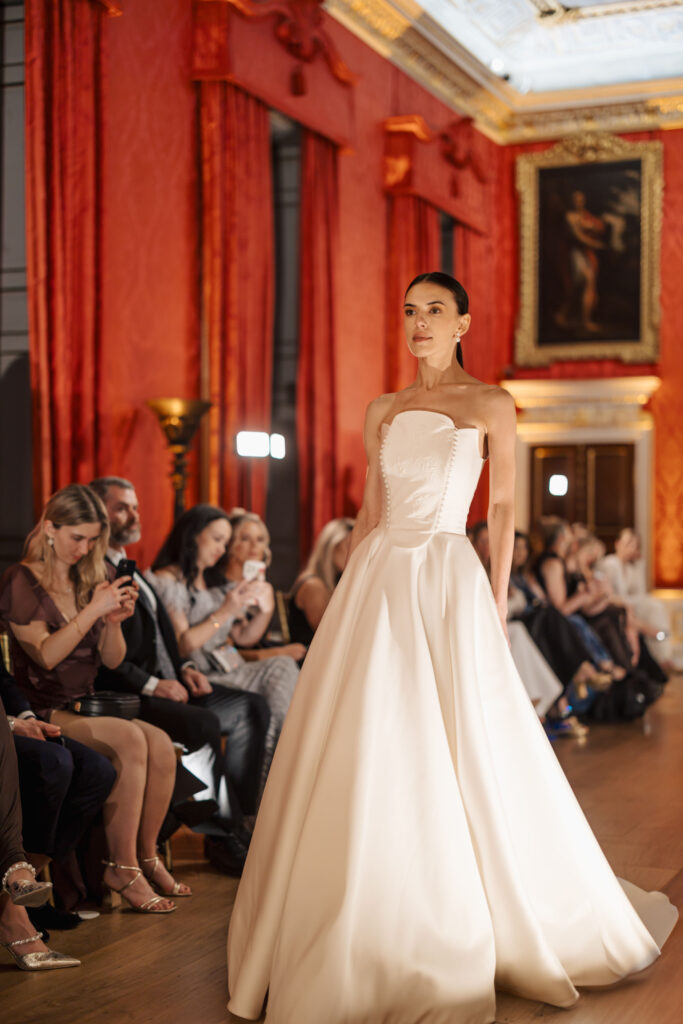 A model walks a runway in an elegant strapless white gown in an ornate room with red walls, gold accents, and a seated audience watching and taking photos.