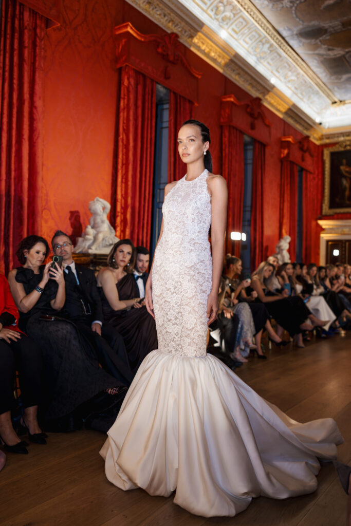 A model walks the runway in an elegant, sleeveless white lace mermaid gown, with a flared skirt, during a fashion show in an ornate, red-walled hall filled with seated guests.