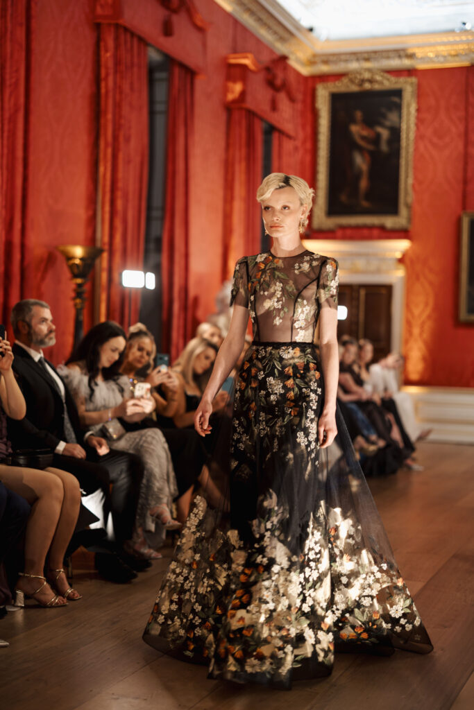 A model walks down a runway in an ornate room with red walls, wearing a sheer black gown decorated with gold and white floral patterns. Seated spectators watch and take photos as she passes.