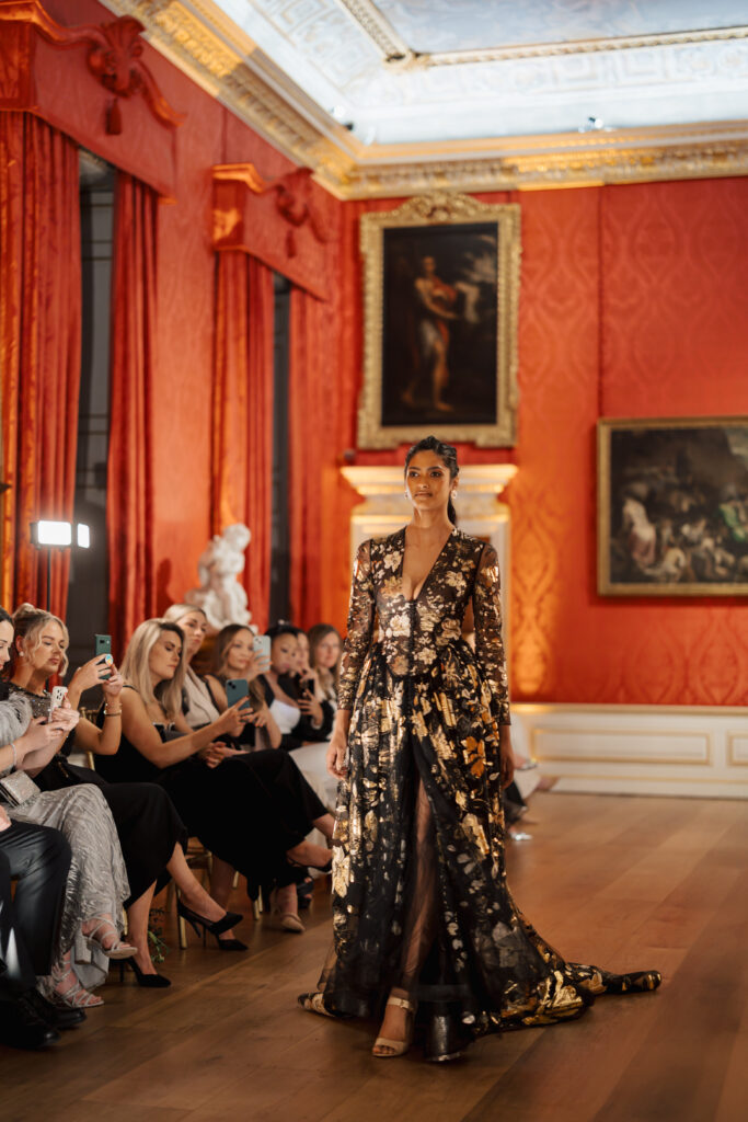 A model walks down a runway in a luxurious room with red walls and gold accents, wearing a long, dark sheer gown with gold patterns. Seated audience members watch and take photos on their phones. Paintings and statues decorate the background.