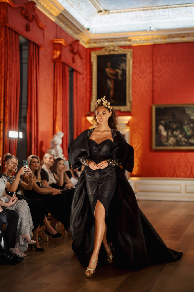 A model in a dramatic black gown and gold headpiece walks a runway in an opulent room with red walls, gold accents, and classical paintings, while an audience watches.