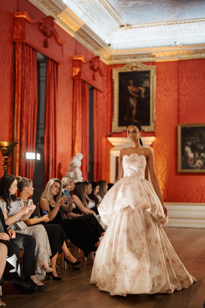 A model walks a runway in an elegant room with red walls, wearing a strapless, floral ballgown with a voluminous skirt. Audience members watch and take photos as art and sculptures decorate the luxurious setting.