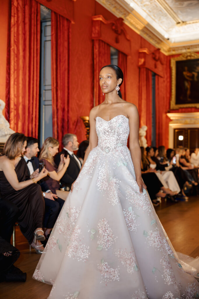 A model walks on a runway in a grand room wearing a strapless, white ballgown adorned with delicate floral details, while an audience sits along the sides watching the fashion show.