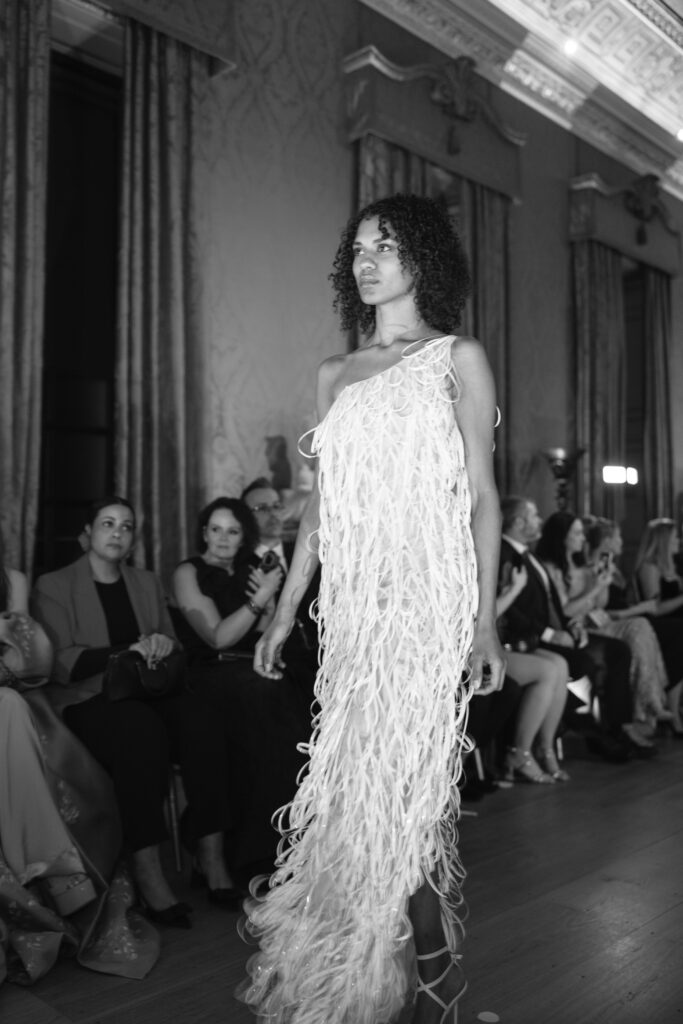 A model with curly hair walks down a runway wearing a textured, fringed dress, while an audience watches attentively in a grand, ornate room. The photo is in black and white.