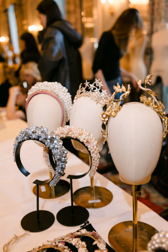 A display of ornate headbands and crowns adorned with pearls and gemstones on stands, with blurred people in the background at a boutique or fashion event.