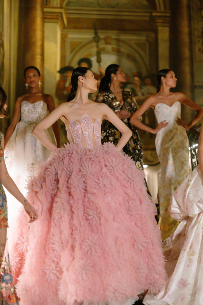 A group of models in elegant evening gowns pose on a grand staircase in an ornate hall. The central model wears a voluminous pink dress adorned with 3D floral details, while others wear gowns in various soft colors and patterns by House of Savin