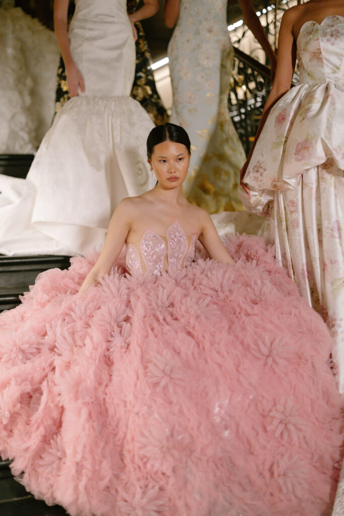 A model sits on stairs wearing a voluminous strapless pink gown adorned with textured floral details. Other models in elegant dresses stand nearby, partially visible in the background.