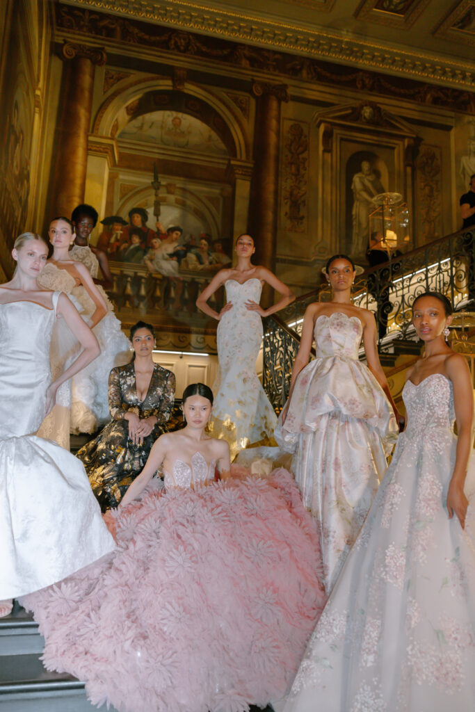 Eight models in elegant, ornate gowns pose on a grand staircase with elaborate, classical murals and columns in the background, creating a luxurious, high-fashion atmosphere.