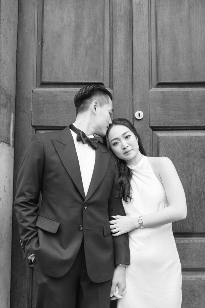 A man in a suit gently kisses the head of a woman in a sleeveless dress as she leans on his shoulder, standing together in front of large wooden doors. The image is in black and white.