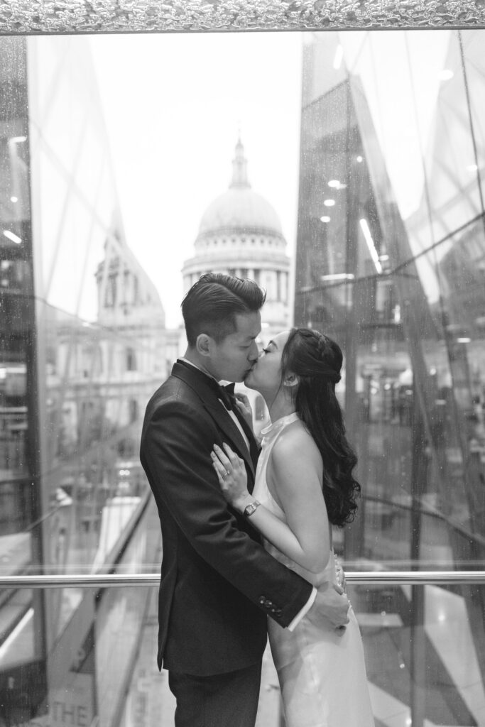 A bride and groom share a kiss indoors, framed by large glass windows. St. Paul’s Cathedral is visible in the background, creating a romantic cityscape scene.