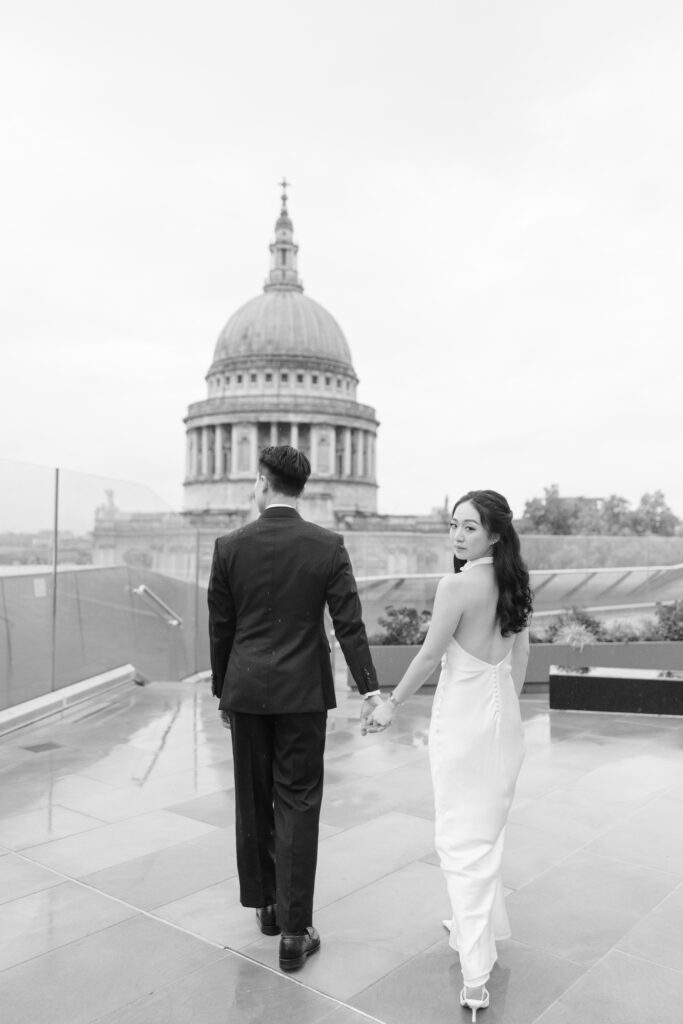 A couple dressed formally walks hand in hand on a rooftop, with St. Paul’s Cathedral in the background. The woman, in a white dress, looks back over her shoulder while the man faces forward. The scene is in black and white.