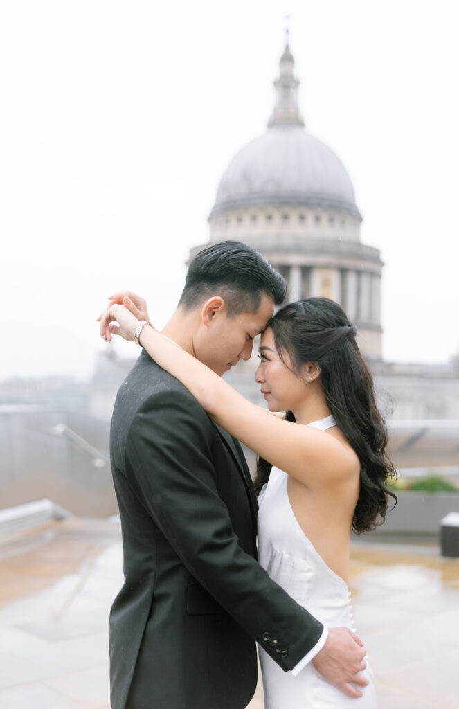 A couple embraces on a rooftop, gazing into each others eyes. The woman wears a white dress, the man a black suit. A large domed building, possibly St. Paul’s Cathedral in London, is blurred in the background.