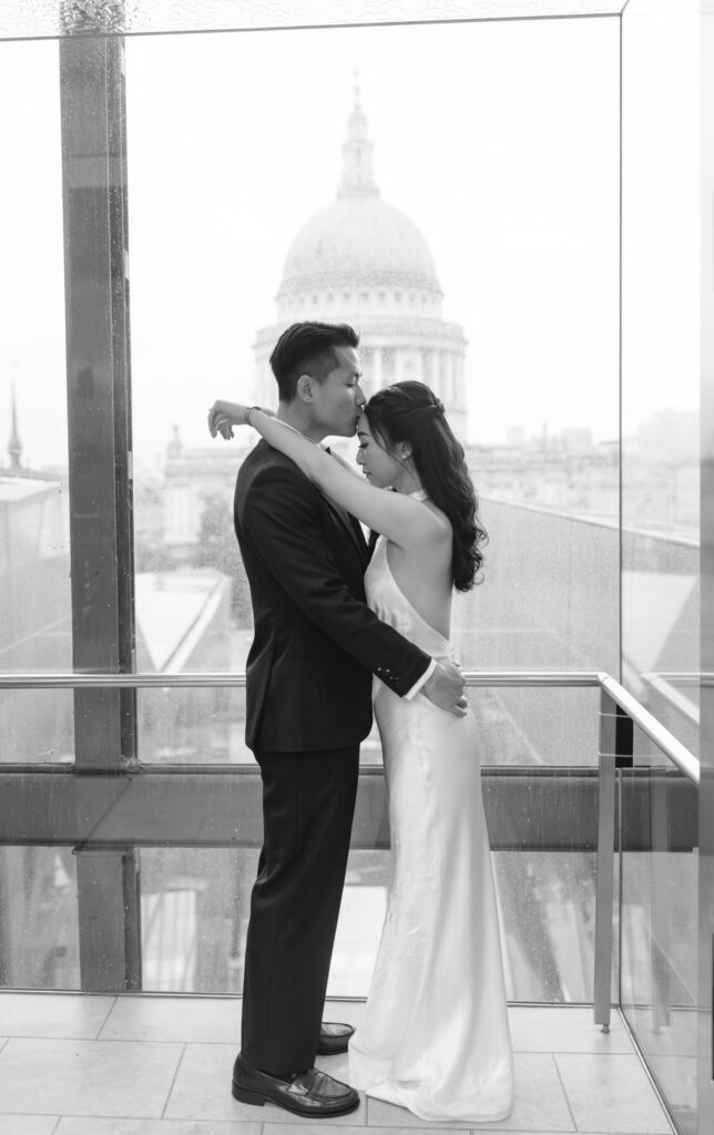 A couple stands embracing in front of a large window with a domed building in the background. The woman wears a white dress, and the man is in a dark suit. The scene is in black and white.