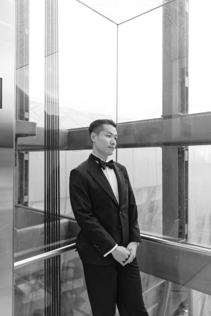 A man in a black tuxedo and bow tie stands in a glass elevator, looking to the side with a calm expression. The elevator is modern with large glass windows and metal frames. The photo is in black and white.