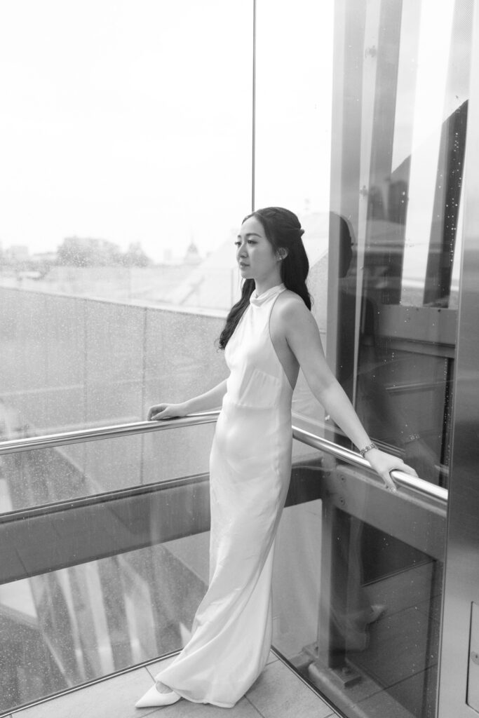A woman in a sleeveless, floor-length white dress stands in a glass elevator, looking out with a calm expression. The scene is in black and white, with city buildings visible through the window.