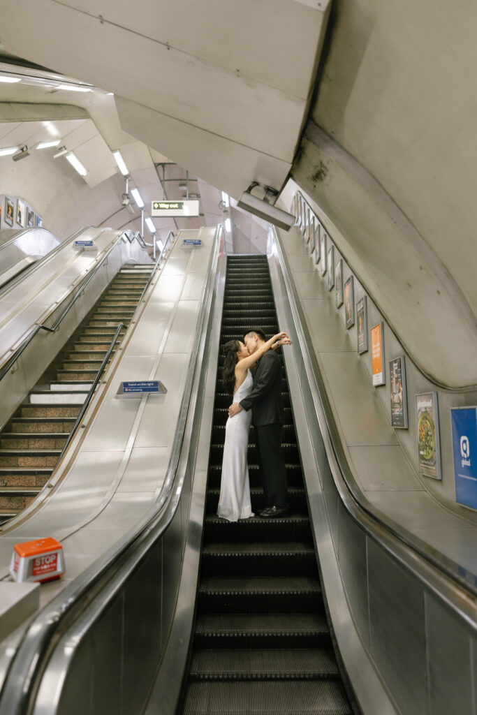 A couple dressed in formal attire shares a kiss while standing on a stationary escalator in an empty subway station, surrounded by metal escalators and advertising posters.
