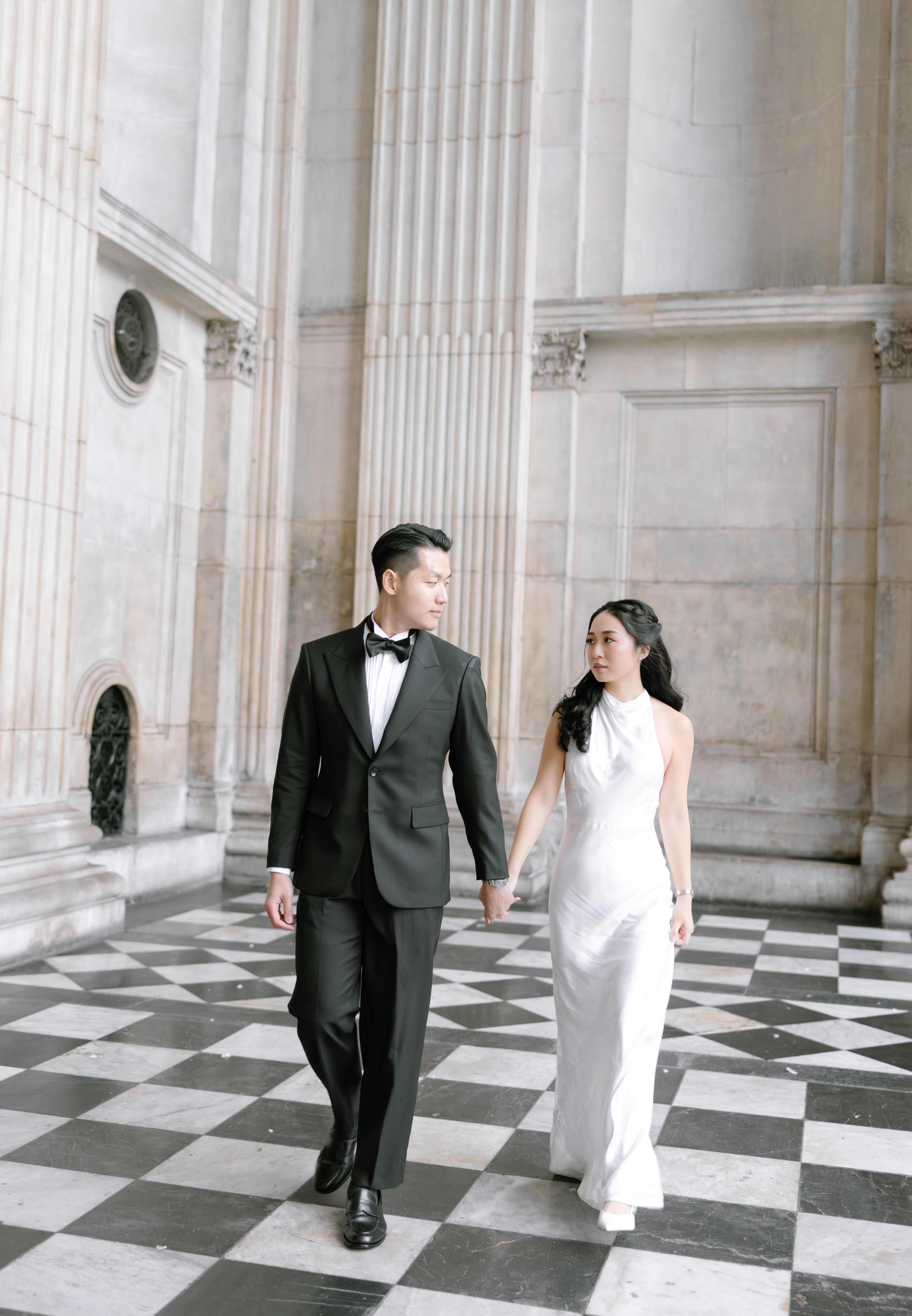 A couple dressed formally—the man in a black suit and bow tie, the woman in a white gown—walk hand in hand on a black-and-white checkered floor in a grand, columned hall.